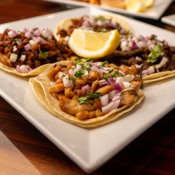 Three delicious Mexican street tacos with various fillings, topped with diced onions, cilantro, and a lemon wedge.